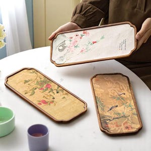 以下が含まれることがあります： 花柄と鳥柄の長方形の木製トレイ。1つはピンクの花と書道、もう1つは赤い果物、3つ目は鳥が描かれています。トレイは白いテーブルの上にあります。