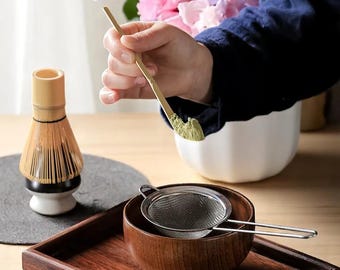 May include: A matcha tea preparation set featuring a bamboo whisk, a wooden bowl, a stainless steel strainer, and a wooden scoop holding green matcha powder. A white ceramic bowl and a wooden tray are also present.