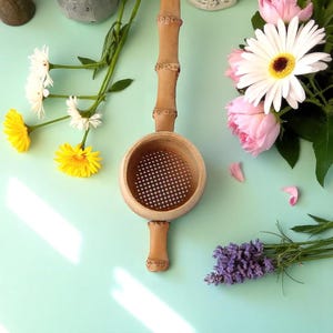 Puede incluir: Un colador de té de bambú hecho a mano con un cuenco redondo perforado y un mango largo. El colador es de color marrón claro y presenta tallas decorativas. La imagen está decorada con flores frescas, incluyendo margaritas blancas, rosas rosas y lavanda.