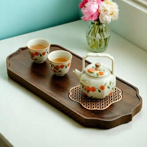 Puede incluir: Una bandeja de té de madera oscura con dos tazas pequeñas y una tetera, todas adornadas con diseños florales naranjas. Un posavasos decorativo de cobre sostiene la tetera. Un jarrón de flores rosas y blancas está en el fondo.