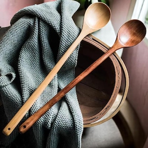 May include: Two wooden spoons with long handles. One spoon is light-colored, the other is a darker brown. The spoons are resting on a gray textured blanket and a wooden basket. The spoons are ideal for cooking and serving.