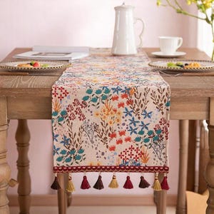 May include: A floral table runner with a cream background and colorful flowers in red, blue, yellow, and green. The runner has a burgundy border with tassels in shades of brown and gold. The runner is placed on a wooden table.