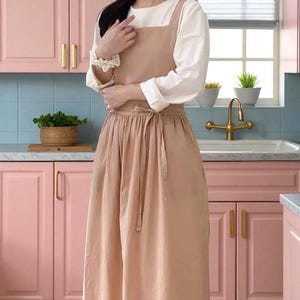 Op de afbeelding: Een beige schort met een lange rok, vastgebonden in de taille, gedragen over een wit shirt met lange mouwen. De schort heeft een slab en bandjes. De achtergrond omvat roze kasten, een blauwe tegelachterwand en een gouden kraan.