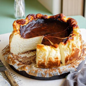 May include: A partially sliced Basque burnt cheesecake on parchment paper. The cake has a dark, caramelized exterior and a creamy, light-colored interior. A knife rests beside the cake, suggesting it's ready to be served. The cake sits on a dark plate.