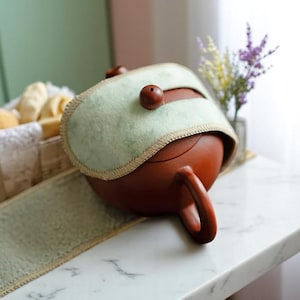 Puede incluir: Una tetera de cerámica marrón con un asa curvada y una tapa verde claro. La tapa tiene una pequeña cuenta de madera en la parte superior y está bordeada con un borde marrón. La tetera está sobre una superficie blanca.