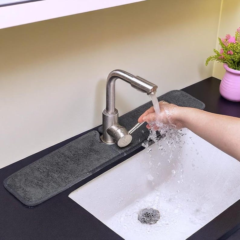 May include: A stainless steel faucet with water flowing into a white sink. A gray absorbent mat sits behind the faucet. A person's hand is under the water stream. A small pink vase with flowers is in the background.
