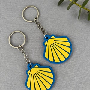 Può includere: Due portachiavi con un design a forma di conchiglia. Ogni conchiglia presenta un centro giallo brillante con creste in rilievo e un bordo blu solido. I ciondoli sono fissati ad anelli in metallo argentato tramite corte catene metalliche.