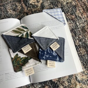 May include: Four fabric bookmarks with a triangular shape. Each bookmark has a different pattern and color, including a blue and white gingham pattern, a green and white floral pattern, a brown and green pattern, and a blue and white pattern. Each bookmark has a small rectangular tag sewn on the bottom with the text "GRACE VALLEY FARM".