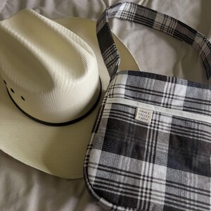 May include: A black and white plaid crossbody bag with a white leather tag that reads "GRACE VALLEY FARM". The bag is next to a white straw cowboy hat.