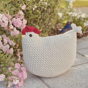 Könnte beinhalten: Ein handgehäkeltes, weißes Häschen mit rotem Kamm und gelbem Schnabel. Das Häschen hat ein schwarzes Auge und steht auf einer Steinoberfläche, mit rosa Blumen im Hintergrund. Dieser Dekorationsartikel ist ideal für die Einrichtung im Landhausstil.