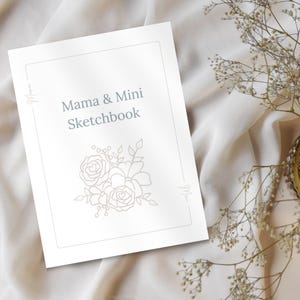 May include: A white sketchbook with the text "Mama & Mini Sketchbook" in a soft gray font. The cover features a floral design and the word "Mama" in script. The sketchbook rests on a white fabric surface.