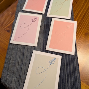 May include: Five greeting cards with paper aeroplane designs. Each card features a different colour scheme: pink, green, and grey. The paper aeroplanes are in various colours, with dotted lines indicating their flight paths. The cards are on a blue fabric.