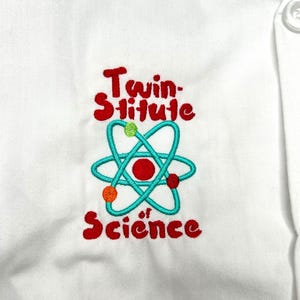 Op de afbeelding: Witte laboratoriumjas met een geborduurd ontwerp. Het ontwerp toont de woorden "Twin-stitute of Science" in het rood, met een turquoise atoom symbool en kleurrijke accenten.