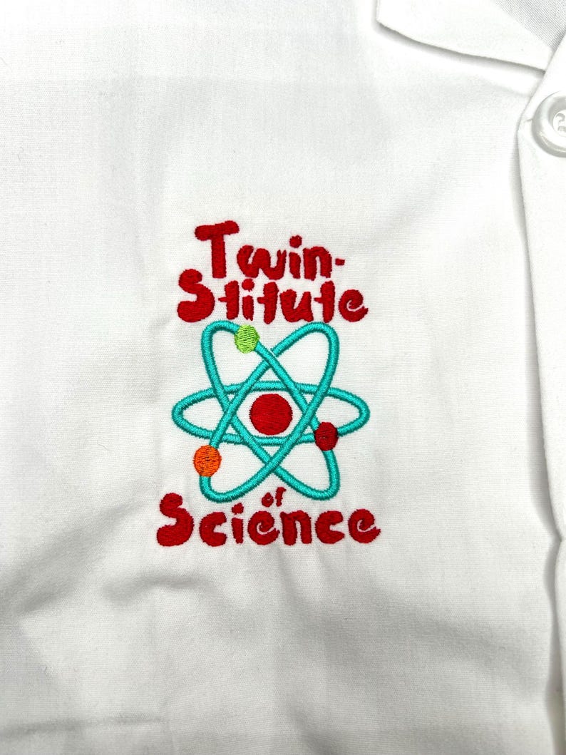 Op de afbeelding: Witte laboratoriumjas met een geborduurd ontwerp. Het ontwerp toont de woorden "Twin-stitute of Science" in het rood, met een turquoise atoom symbool en kleurrijke accenten.