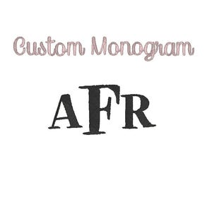 May include: A white background with the text "Custom Monogram" in a light pink script font. Below the text are the black letters "AFR" in a bold, serif font. The letters are slightly textured.