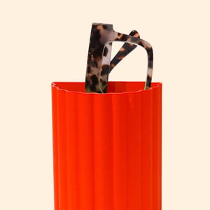 May include: A vibrant orange cylindrical container with a ridged exterior, holding a pair of eyeglasses with a tortoiseshell pattern. The container has a wavy top edge and casts a shadow on the light background.