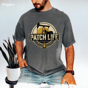 Könnte beinhalten: Ein anthrazitfarbenes Kurzarm-T-Shirt mit einer kreisförmigen Grafik, die eine gelbe Sonne, die schwarze Silhouette einer Ölpumpe und den Text PATCH LIFE in fetten schwarzen Buchstaben zeigt. Darunter befinden sich drei kleine gelbe Öltropfen.
