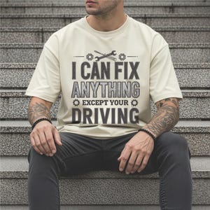 May include: Cream-colored t-shirt with the text "I CAN FIX ANYTHING EXCEPT YOUR DRIVING" in gray. The design includes tools and gears. The person is wearing black jeans and a black bracelet.