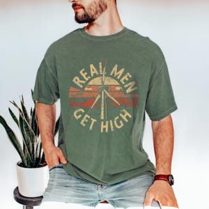 May include: Olive green t-shirt with a distressed graphic. The graphic features a wind turbine design with the words "REAL MEN GET HIGH" in a circular layout, using orange and cream colours. The shirt has short sleeves and a crew neck.
