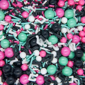 May include: A colorful mix of sprinkles, including pink, black, white, and mint green spheres, rods, and musical note shapes. The sprinkles are scattered, creating a vibrant and textured pattern, perfect for decorating baked goods.