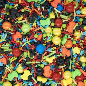May include: A colorful mix of sprinkles and candies, including star shapes, game controllers, and round candies in various colors. The sprinkles are a mix of red, green, yellow, orange, blue, and black.