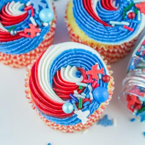 May include: Three red, white, and blue cupcakes with sprinkles and candy. The cupcakes are decorated with a swirl of frosting and are topped with red, white, and blue sprinkles and candy.