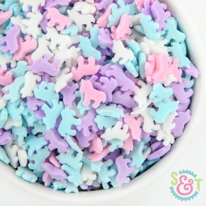 May include: A close-up shot of a white bowl filled with colorful unicorn-shaped sprinkles. The sprinkles are in pastel shades of pink, purple, blue, and white. The image is brightly lit, showcasing the details of the sprinkles.