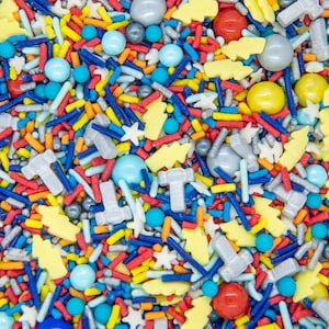May include: A colorful mix of sprinkles for baking and decorating. The sprinkles include stars, small balls, and long, thin sticks in various colors, including red, blue, yellow, and orange. There are also larger, white, and silver sprinkles shaped like hammers and lightning bolts.