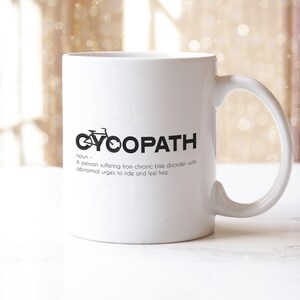 May include: White ceramic mug with the word "CYOOPATH" in black, featuring a bicycle graphic. The mug also includes a definition: "A person suffering from chronic bike disorder with abnormal urges to ride and feel free."