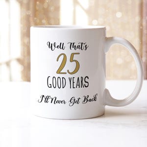May include: White ceramic mug with the text "Well That's 25 Good Years I'll Never Get Back" in black and gold lettering. The mug has a curved handle and is set against a blurred background.