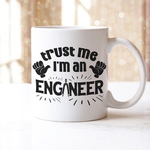 May include: White ceramic mug with a curved handle. The mug features the text "trust me I'm an ENGINEER" in black, along with hand illustrations and a compass design. The mug is designed for engineers.