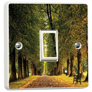 May include: A white light switch cover with a printed image of a tree-lined pathway. The scene depicts a path covered in autumn leaves, a park bench, and a bright, sunny day. The switch is centrally located.