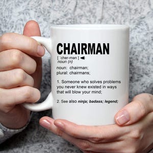 May include: White ceramic mug with the word "CHAIRMAN" in bold black letters. The mug features a definition of the word, including its pronunciation and meaning. The definition includes the phrases "solves problems" and "blow your mind."