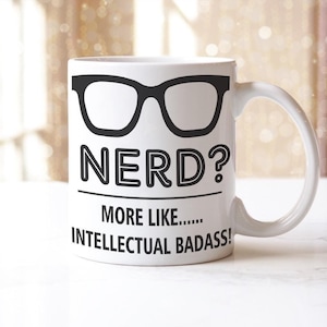 May include: White ceramic mug featuring a black glasses graphic and the text "NERD? MORE LIKE... INTELLECTUAL BADASS!" The mug has a curved handle and is suitable for hot or cold drinks.