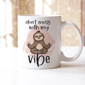May include: White ceramic mug with a cartoon sloth meditating. The mug has the text "don't mess with my vibe" in black script. A pink watercolor-style background is behind the sloth. The mug has a curved handle.