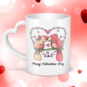 Puede incluir: Taza de cerámica blanca con asa en forma de corazón. La taza presenta una colorida ilustración de dos pájaros posados en una rama, rodeados de corazones y elementos florales. El texto "Happy Valentine Day" está impreso debajo.