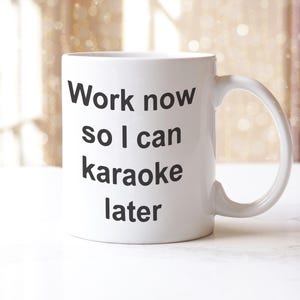 May include: White ceramic mug with the text "Work now so I can karaoke later" in black. The mug has a curved handle. The background is blurred. A fun, motivational coffee mug for the office or home.