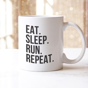 May include: White ceramic mug featuring the text "EAT. SLEEP. RUN. REPEAT." in black, bold lettering. The mug has a curved handle and is set against a blurred background.
