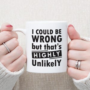 May include: White ceramic mug with the text "I COULD BE WRONG but that's HIGHLY Unlikely" in black. The mug is held by hands wearing rings, against a neutral background. A humorous gift idea.