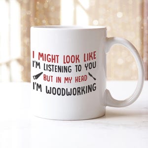 May include: White ceramic mug with a curved handle. The mug features the text "I MIGHT LOOK LIKE I'M LISTENING TO YOU BUT IN MY HEAD I'M WOODWORKING" in red and black, with saw and chisel graphics.
