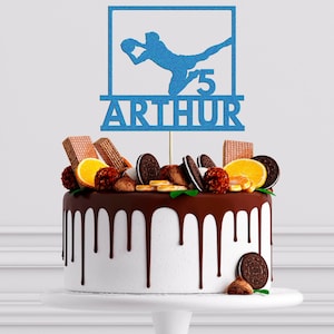 May include: Blue glitter cake topper featuring a silhouette of a person playing rugby, the number 5 and the name Arthur. The topper is on a chocolate cake decorated with orange slices, chocolate chips and other decorations.