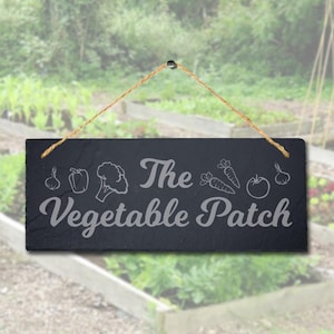 May include: A rectangular black slate sign with the words "The Vegetable Patch" in a decorative font. The sign features etched illustrations of vegetables, including a pepper, broccoli, carrots, and a tomato. A twine loop is attached for hanging.