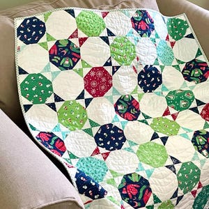 May include: A white quilt with a geometric pattern of green, blue, and red octagons and stars. The quilt is made with a variety of fabrics featuring winter holiday themes, including snowmen, snowflakes, and reindeer.