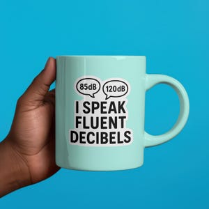 Puede incluir: Una taza de cer&aacute;mica azul claro con un asa blanca. La taza presenta el texto "I SPEAK FLUENT DECIBELS" en negro, con dos bocadillos encima, cada uno con un nivel de decibelios. El fondo es azul liso.