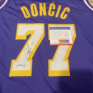 May include: Purple basketball jersey with the name "DONCIC" in yellow block letters and the number "77". The jersey has white trim and an NBA logo. A certificate of authenticity is attached.