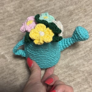 Könnte beinhalten: Eine türkisfarbene Häkel-Gießkanne mit Griff und Ausguss, gefüllt mit bunten Häkelblumen in Gelb-, Rosa-, Weiß- und Grüntönen. Der handgefertigte Artikel wird in einer Hand gehalten.