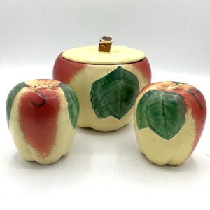 Op de afbeelding: Een keramische keuken set in de vorm van een appel, inclusief een pot met deksel en twee zout- en peperstrooiers. De set heeft een rood-crème kleurenschema met groene bladaccenten. De pot heeft een bruine stelen handvat.