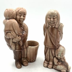Peut inclure: Deux figurines en céramique émaillée marron. La première représente une personne portant un enfant à côté d'un récipient cylindrique en forme de panier. La seconde montre une personne debout avec un petit enfant assis à ses pieds. Finition brillante.