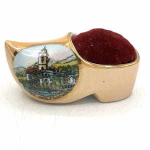 May include: A ceramic clog-shaped pincushion with a glossy cream finish and gold trim. The side features a colorful illustration of a town with a church tower and the text "Weißes Rössl ST. WOLFGANG". A deep red velvet cushion fills the opening.