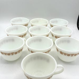 May include: A set of ten white milk glass cups arranged in rows. Each cup features a repeating brown floral pattern around the exterior base. The cups have a rounded shape with a small handle on the side, showcasing a consistent design across all pieces.